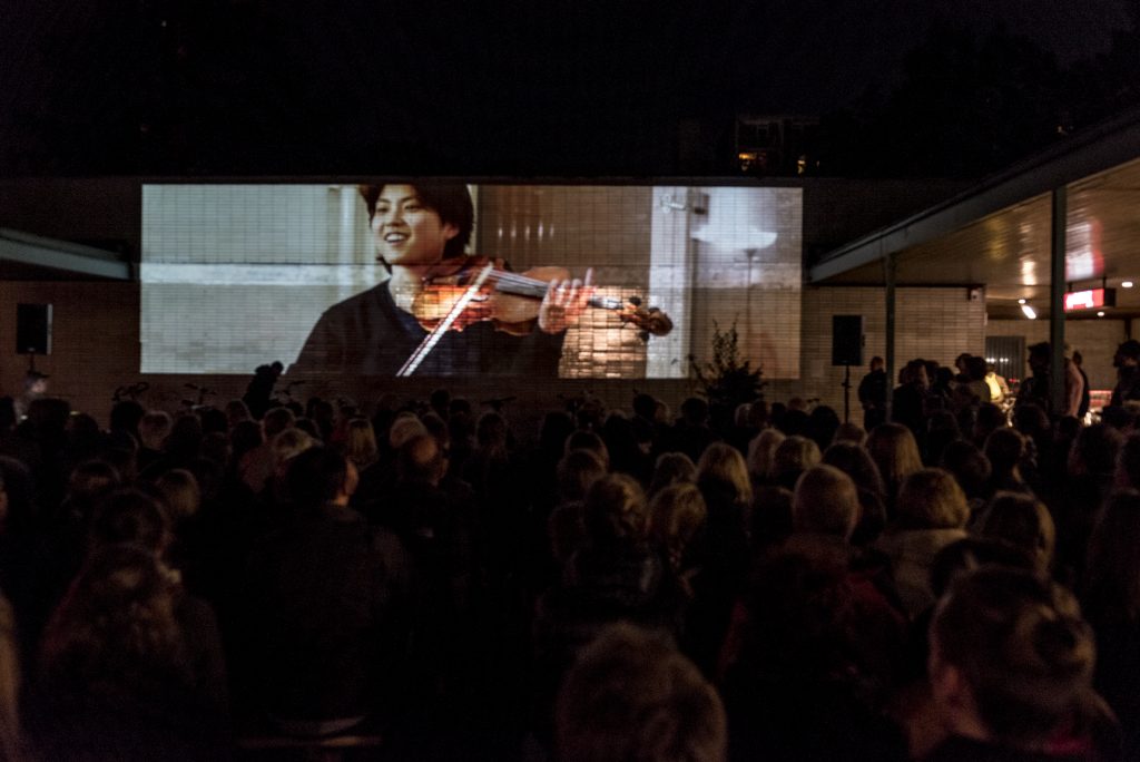 Menschen sitzen im Dunkeln auf einem Platz, an eine Wand wird eine Frau, die Geige spielt projeziert