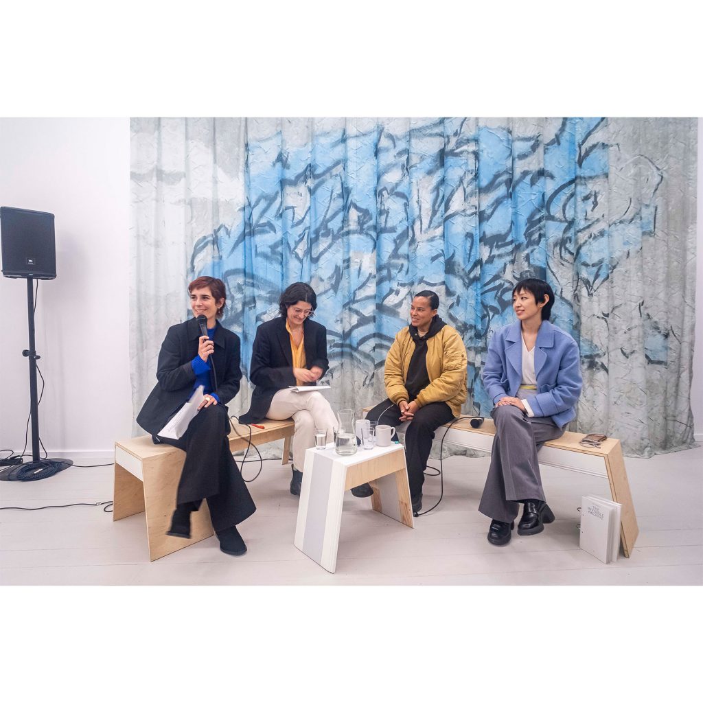 Four people on chairs in front of an artwork, one speaks into a microphone