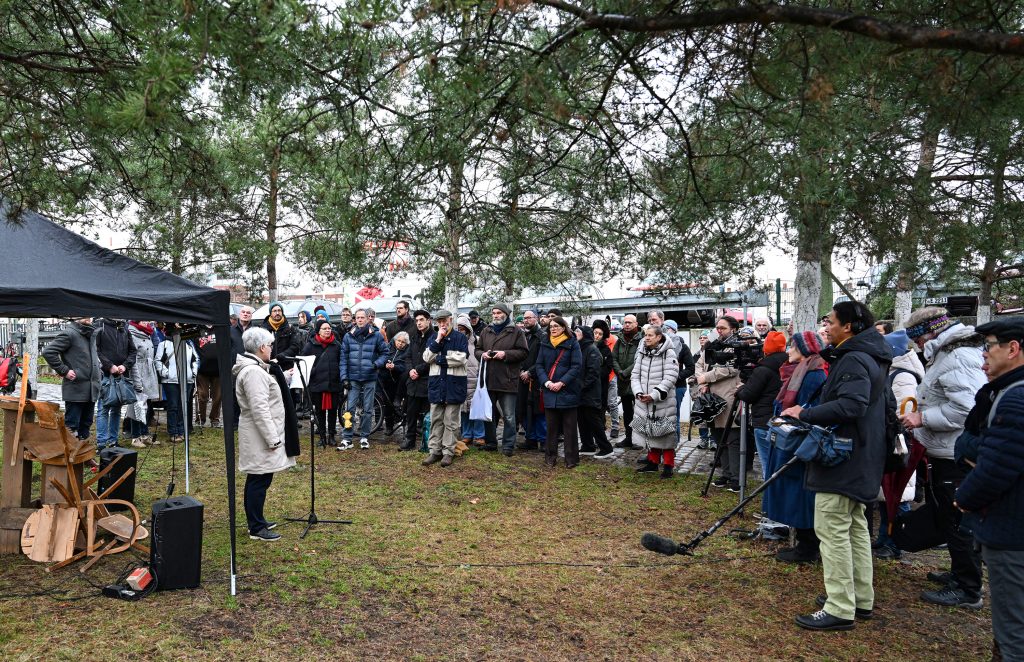 Menschen unter Nadelbäumen hören einer Frau am Mikrofon zu
