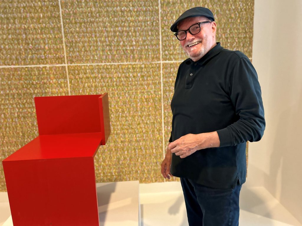 Lächelnder Mann mit Brille und Schiebermütze neben rotem Stuhl vor goldenem Hintergrund