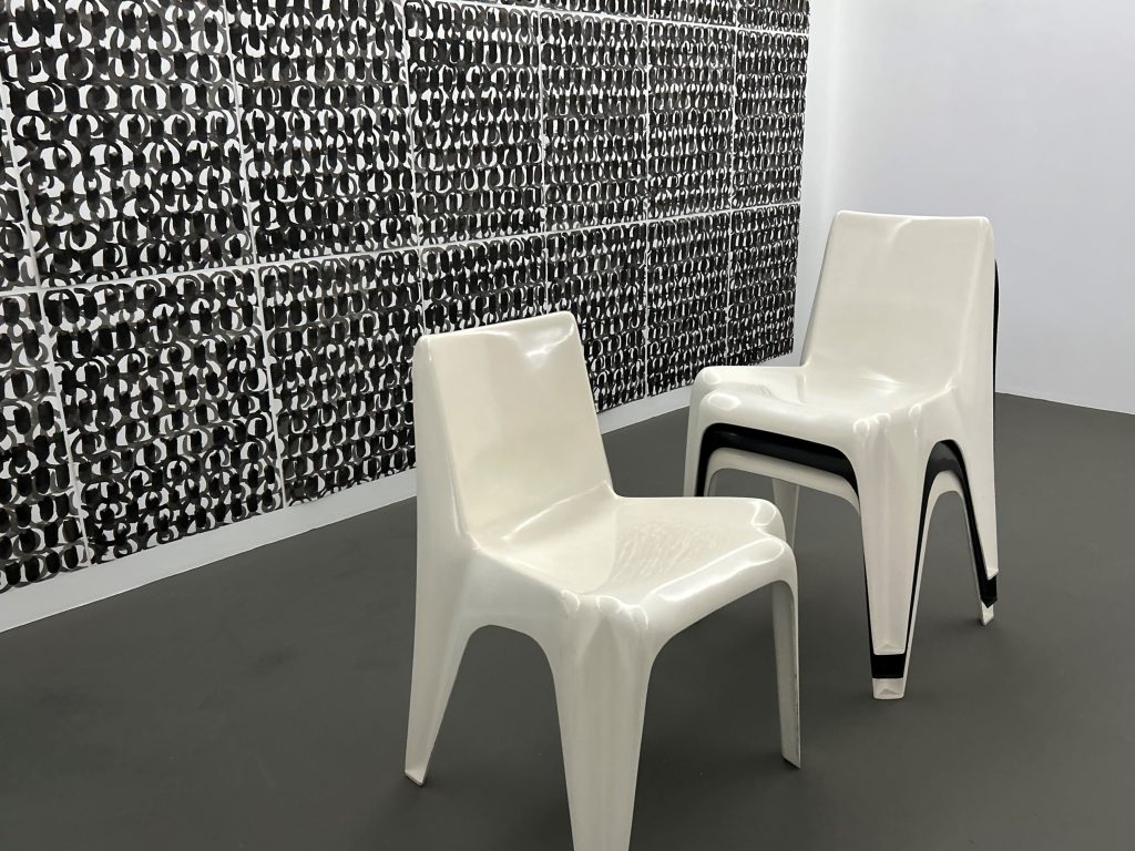 Einzelner Stuhl neben drei gestapelten Stühlen. Im Hintergrund schwarz-weiß gemusterte Tapete