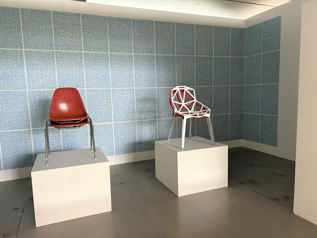 Zwei Sockel, auf einem gestapelte rundliche Stühle auf dem anderen Stuhl aus geometrischen Formen. Im Hintergrund blassblaue Tapete