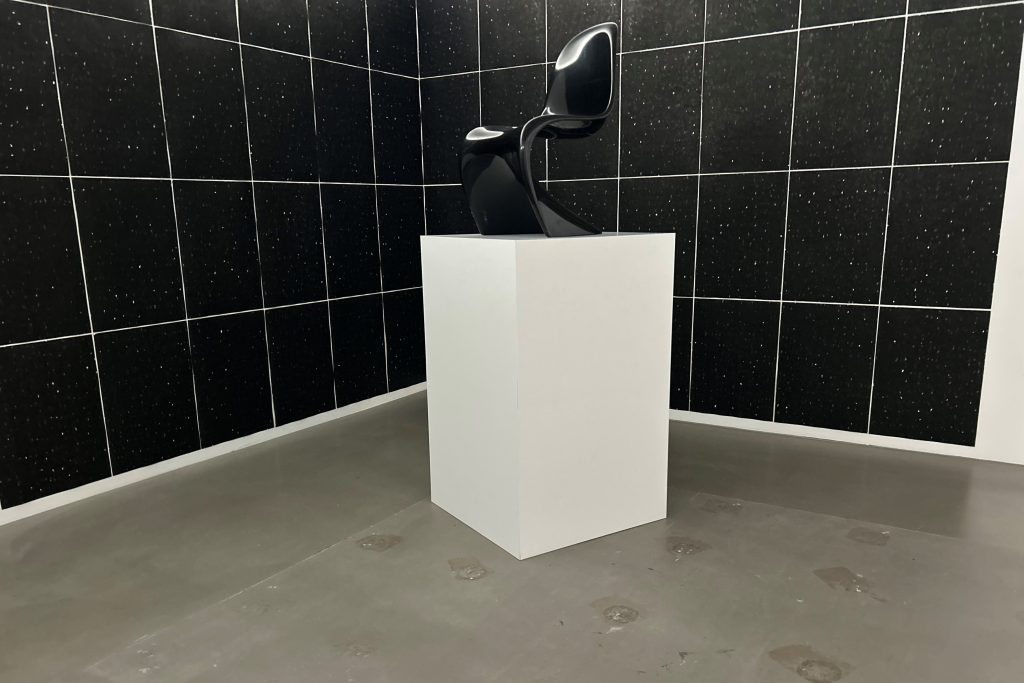 Futuristischer schwarzer Stuhl auf Sockel. Im Hintergrund schwarze quadratische Tapete