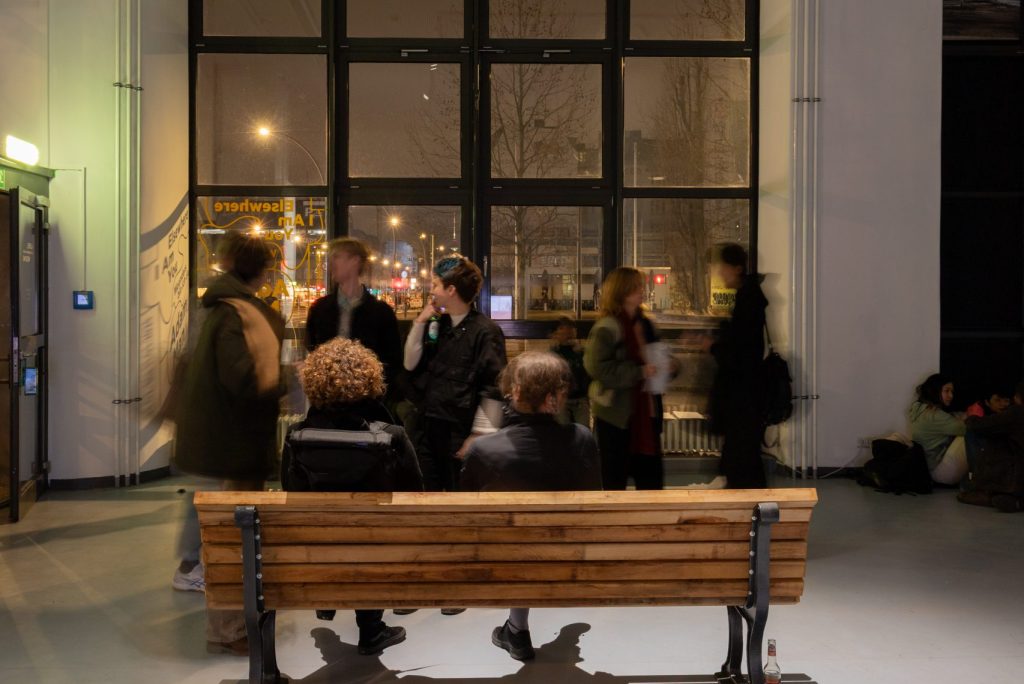 Parkbank mit Menschen darauf und dahinter in der Galerie Wedding