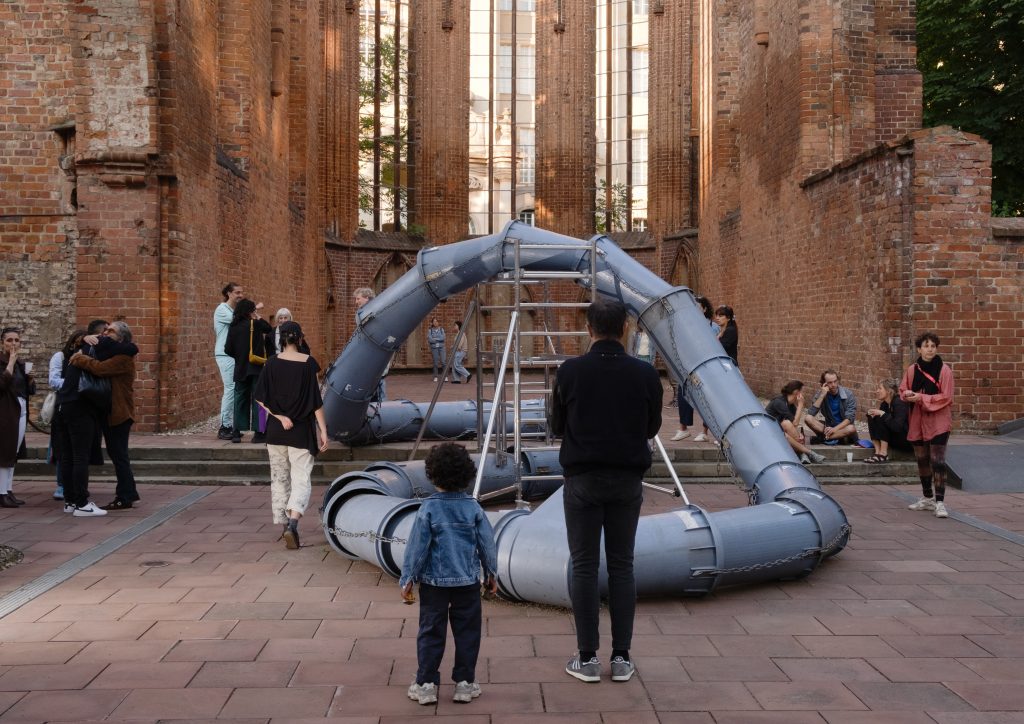 Verschlungene Rohre in einer Kirchenruine, darum herum die Skulptur betrachtende Menschen