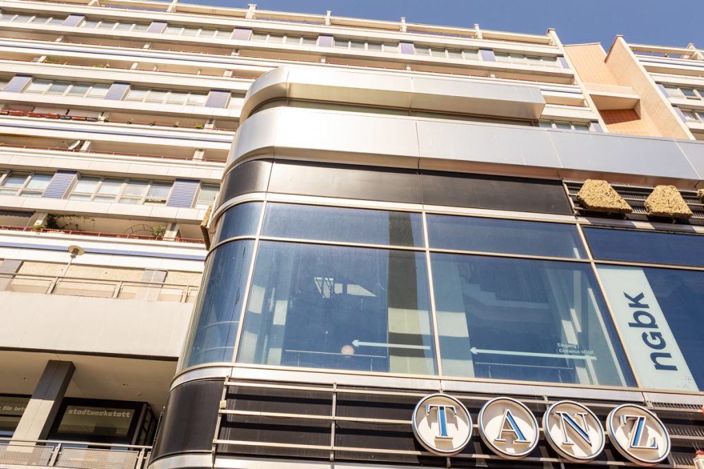 Mehstöckige Betonfassade mit Balkonen und einem auskragenden, verglasten Gebäudeteil im Vordergrund.