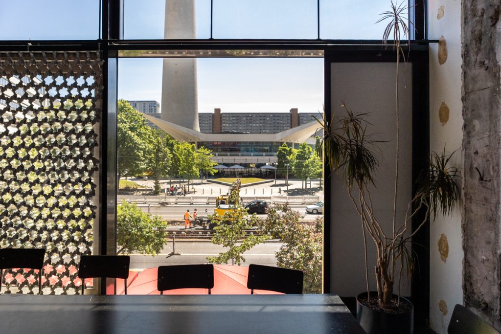 Im Vordergund ein Besprechungstisch mit Stühlen, der Blick geht durch ein bodentiefes Fenster auf einen sonnigen Platz.