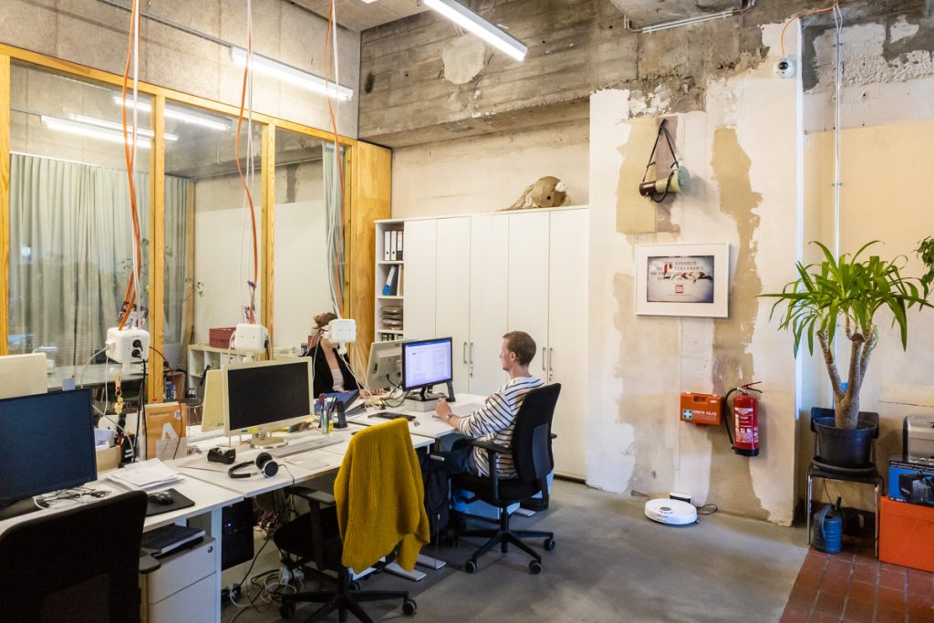 Ein offener Büroraum mit Computerarbeitsplätzen, an denen zwei Menschen sitzen, unverputzete Wände.