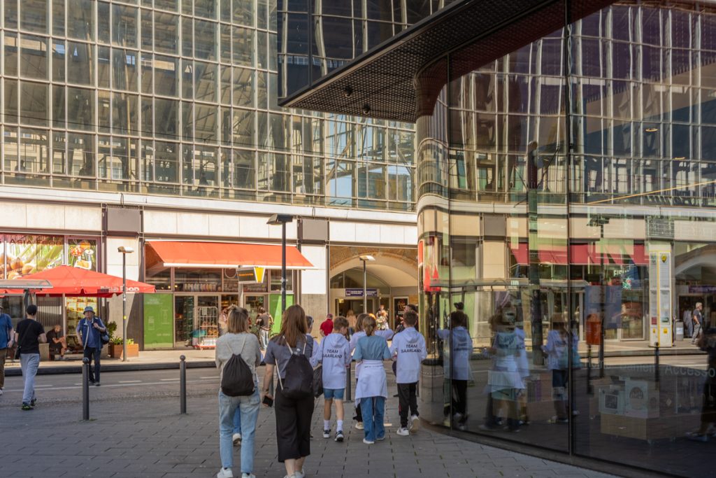 Im Vordergrund geht eine Gruppe Kinder und Erwachsene an einer Glasfassade vorbei, im Hintergrund ist ein ebenfalls verglastes Bahnhofsgebäude zu sehen.