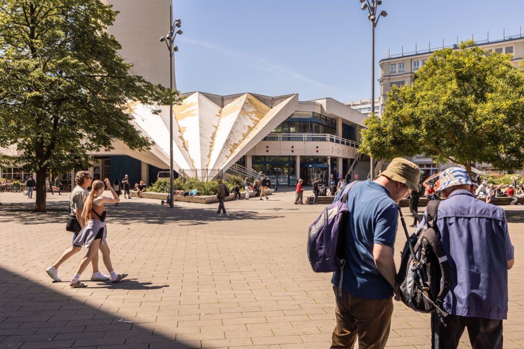 Ein gepflasterter Platz mit Menschengruppen im Sonnenlicht, rechts vorne zwei Männer mit Rucksäcken, Wanderstock und Fischerhüten.