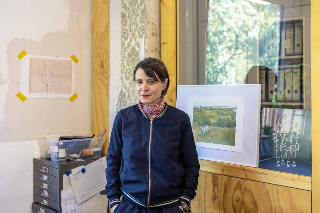 Eine Frau mit dunklen Harre, dunkelblauer Collegejacke und Halstuch steht in einem Büro und blickt in die Kamera.