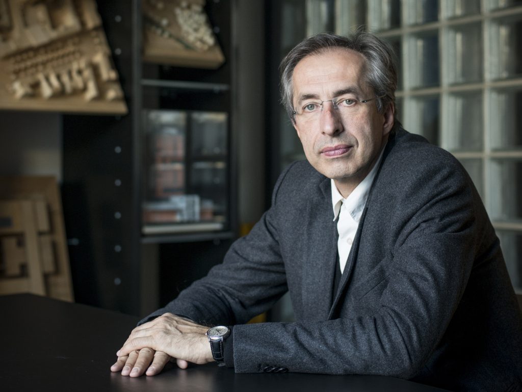 Mann mit Brille, grauem Haar, dunkelgrauem Anzug, weißem Hemd ohne Krawatte an einem dunkelbraunen Tisch.