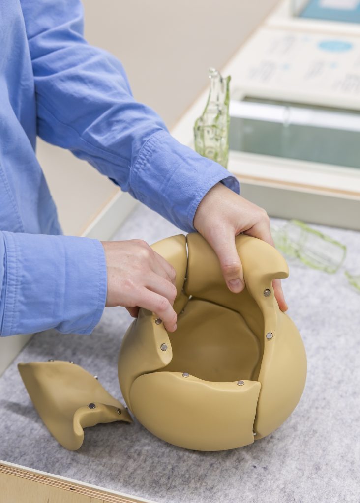 Mensch in hellblauem Hemd, nur die Arme sind zu sehen, setzt eine braune Vase aus einzelnen Fragmenten zusammen.