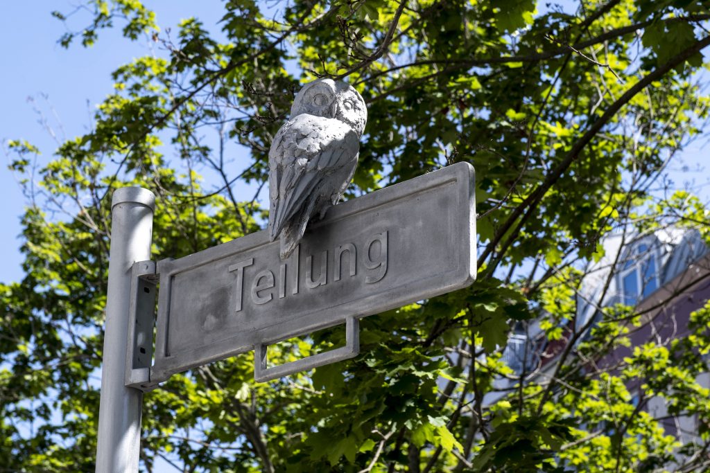 An owl made of metal sits on a street sign that says Teilung