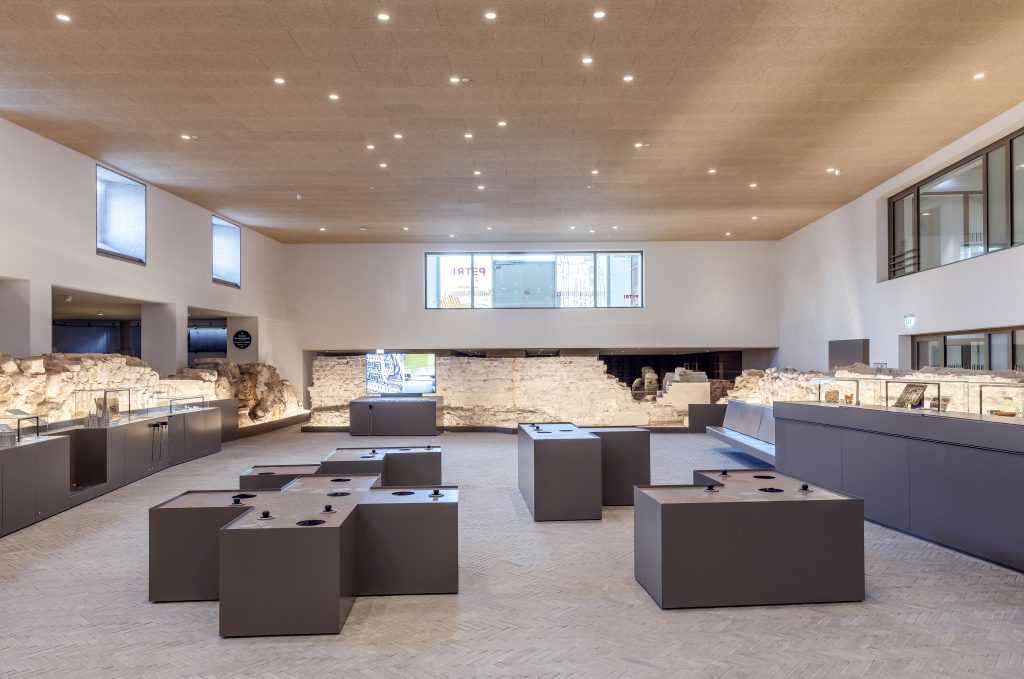 Ein weiter, zweistöckiger Ausstellungsraum, im oberen Geschoss Fenster. Eingerahmt wird er an allen Seiten von hellgrauen Steinfundamenten, davor dunkelgraue Blöcke mit Infotafeln, Vitrinen und Stationen mit Joysticks.