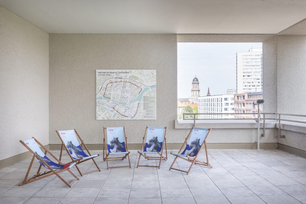 Ein weiträumiger Balkon, fünf Liegestühle, aus Holz und Stoff, mit Bärenmotiv, an der Brüstung ein Fernglas, im Hintergrund die Skyline einer Stadt.