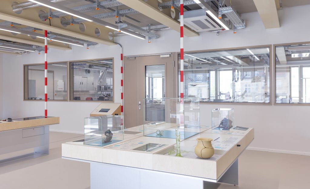 Heller Laborraum, im Vordergrund ein großer Tisch mit Vitrinen, Infotafeln und Objekten zum Anfassen, im Hintergund durch eine Glaswand abgetrennt das eigentliche Labor.
