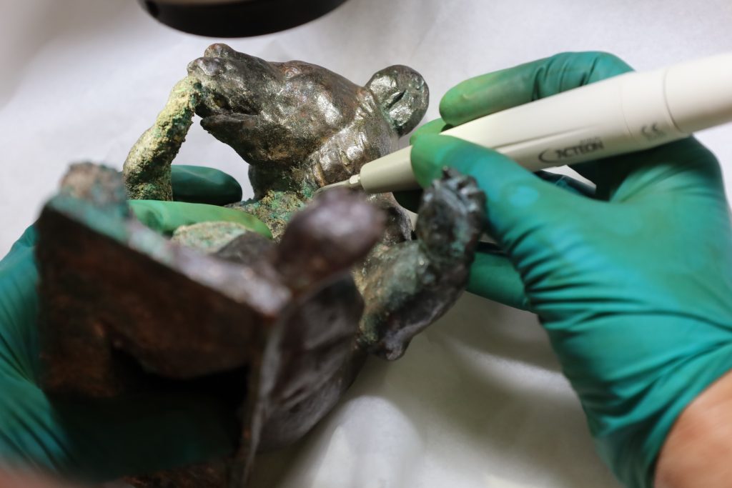 Eine Hand mit grünen Handschuhen bearbeitet mit einem stiftartigen elektrischen Gerät eine Bärenfigur mit grüner Patina.