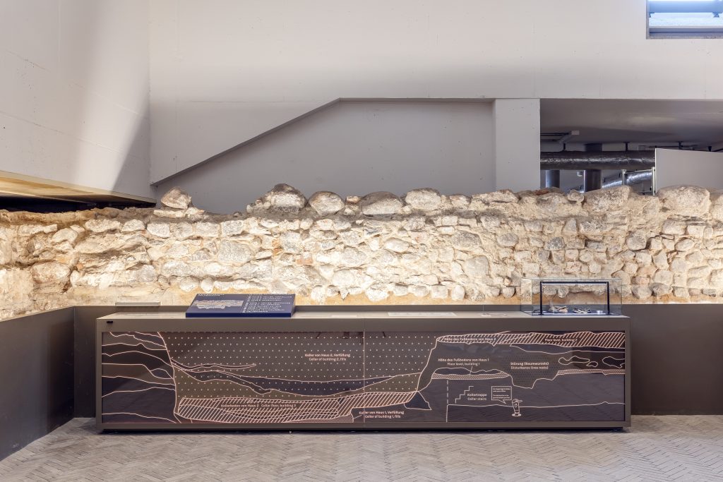 Reste einer hellgrauen Steinmauer, davor in Dunkelgrau eine Infotafel, die eine Vielzahl übereinanderliegender Bodenschichten nachzeichnet.