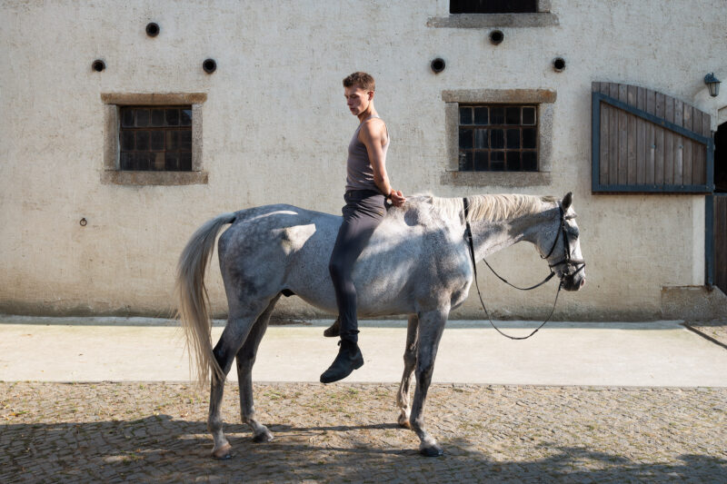 Mensch sitzt mit hinter dem Rücken verschränkten Armen rückwärts auf einem Pferd.
