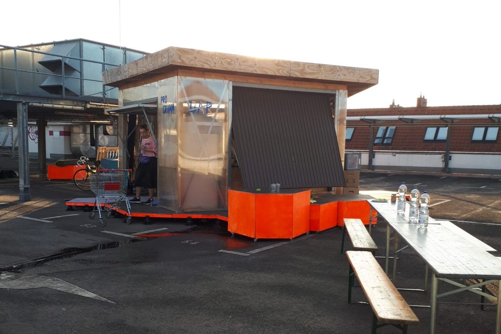 Ein aus Holz- und Plexiglas zusammengebauter Schuppen auf einem Dach, davor Bierbänke.