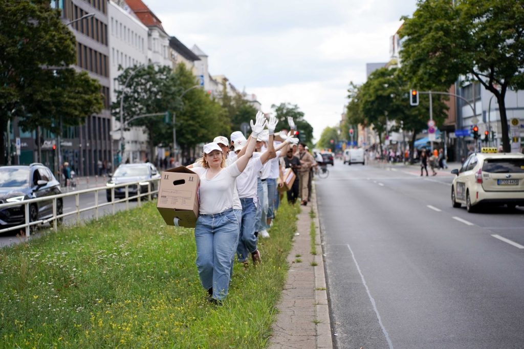 Eine Reihe Menschen läuft hintereinander auf dem grasbewachsenen Mittelstreifen einer mehrspurigen Straße. Sie haben weiße T-Shirts an, tragen einen Umzugskarton in einer Hand und winken mit der anderen.