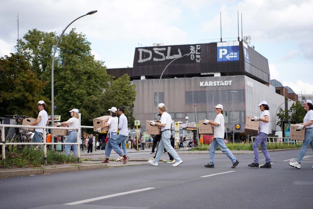 Menschen in Jeans und weißem Oberteil überqueren eine Straße mit Umzugskartons in der Hand. Im Hintergrund ist ein Kaufhaus (Karstadt) zu sehen.