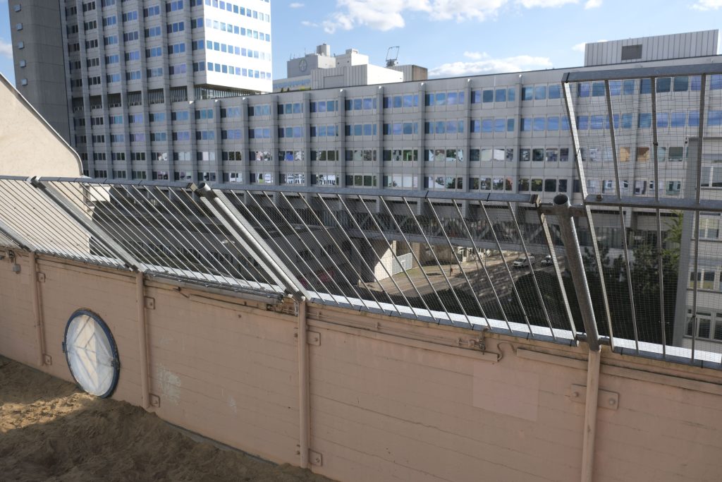 Eine Dach-Brüstung mit Bullauge zum Durchschauen, im Hintergrund ein Hochhauskomplex.