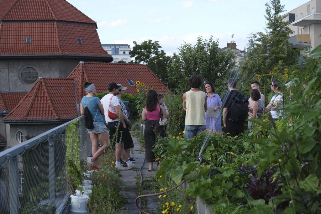 Eine Gruppe Menschen auf einem üppig bewachsenen Dachgarten, im Hintergrund ein neobarockes Gebäude.