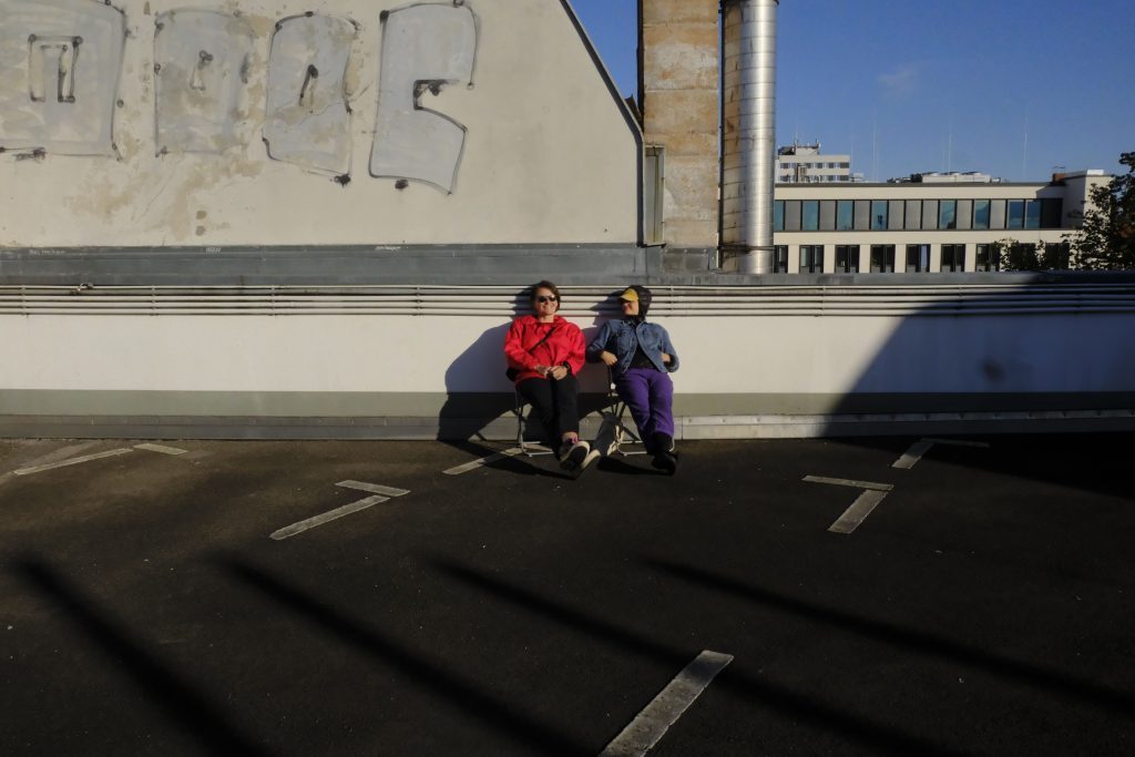 Zwei Personen sitzen auf einem Parkdeck an eine Brüstung gelehnt in der Sonne, dahinter ein Dachgiebel.