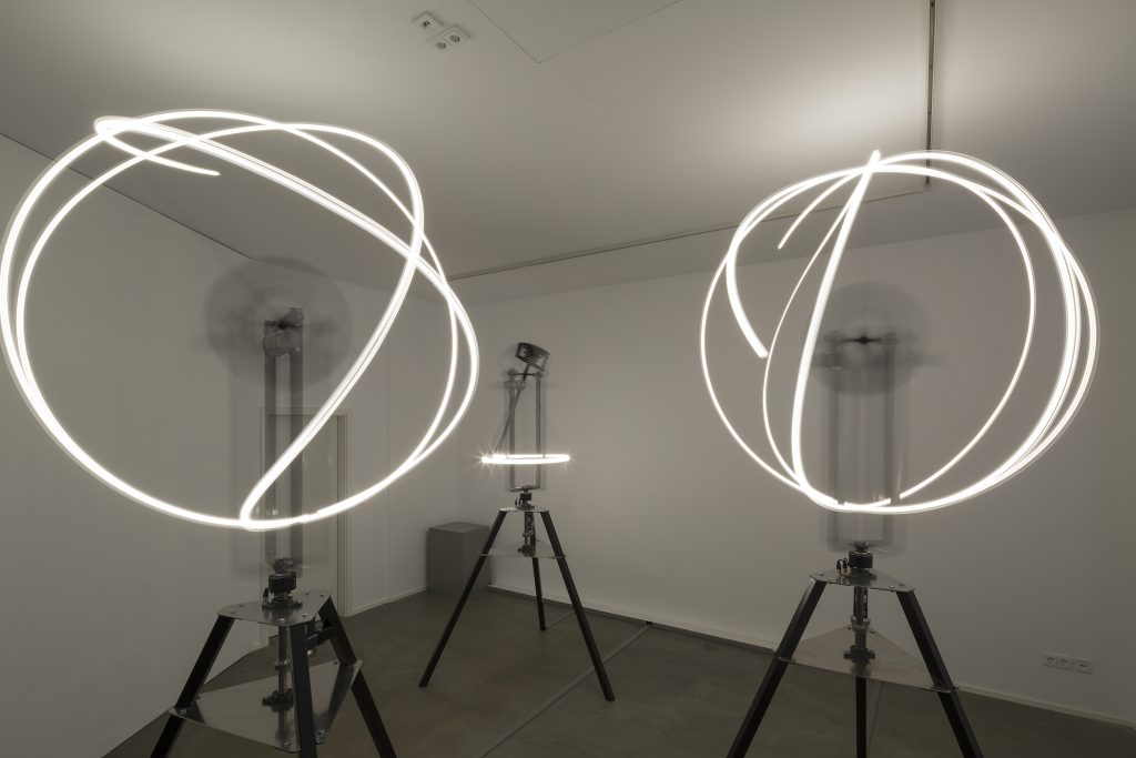 Drei Skulpturen aus weiß-leuchtenden Neonröhren, die auf einem dreibeinigen Gestell montiert sind.