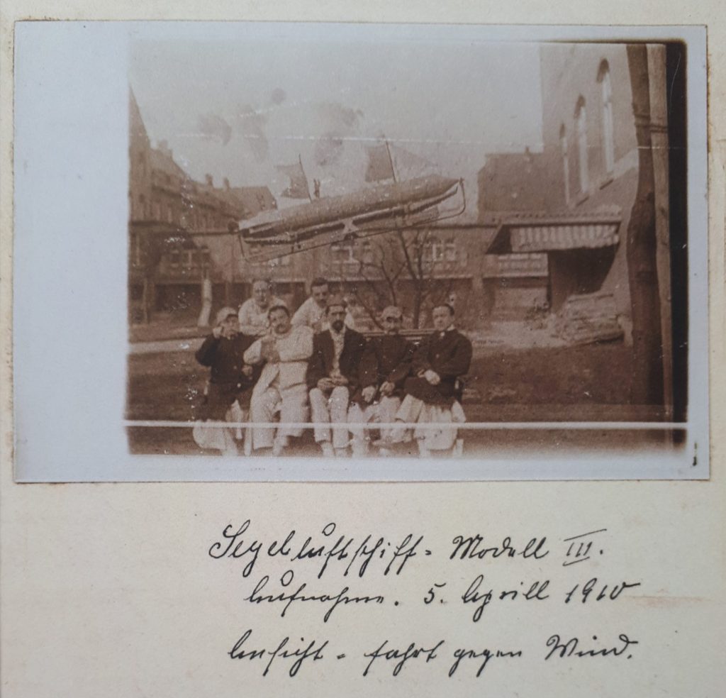 Schwarzweißfotografie: Eine Gruppe von Männern sitzt dichtgedrängt auf einer Bank in einem Innenhof, hinter ihnen scheint ein Luftschiff mit Segeln zu schweben.