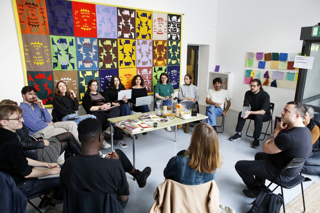 Gruppe im Stuhlkreis in Raum mit bunten Stoffen an der Wand