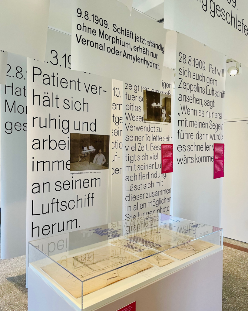 Im Vordergund ein Schaukasten mit Zeichnungen, dahinter dichtgehängt mehrere weiße Papierfahnen mit Text und Bild.