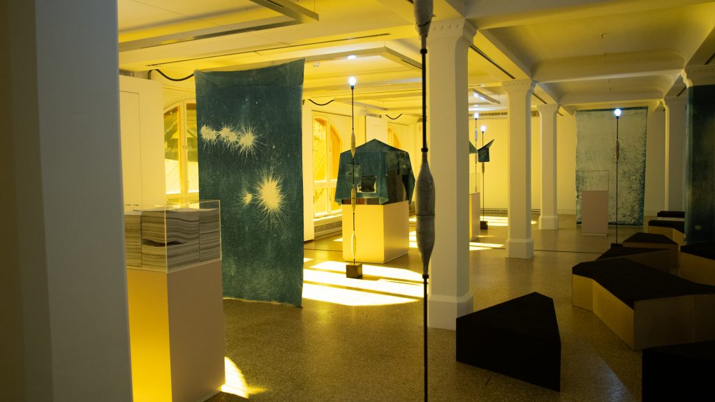 In gelbes Licht getauchter Ausstellungsraum mit verschiedenen Exponaten, Stehlampen und Sitzgelegenheiten.