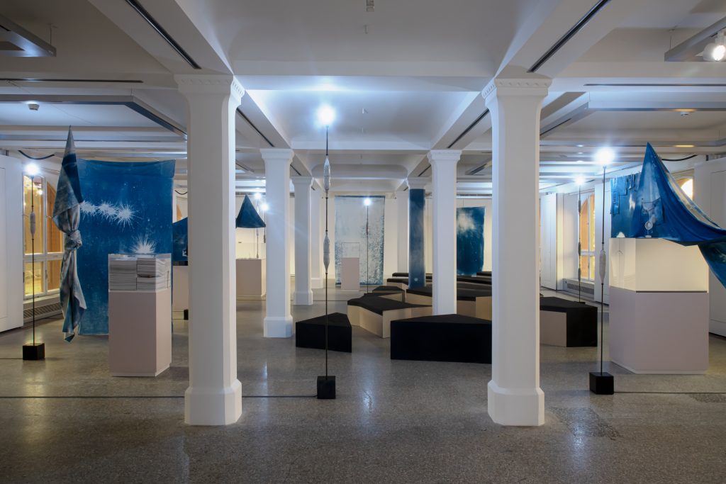 Heller Ausstellungsraum mit weißgestrichenen Stützen, diversen Vitrinen, blauen Stoffbahnen, schlanken, hohen Stehlampen sowie einer Vielzahl an schwarzbezogenen Sitzgelegenheiten.