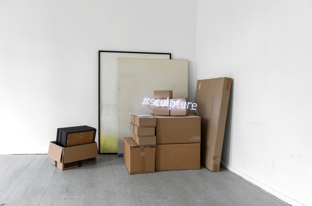 Mehrere teils gestapelte Pappkartons und Bilderrahmen stehen in der Ecke eines weißgestrichenen Raums.