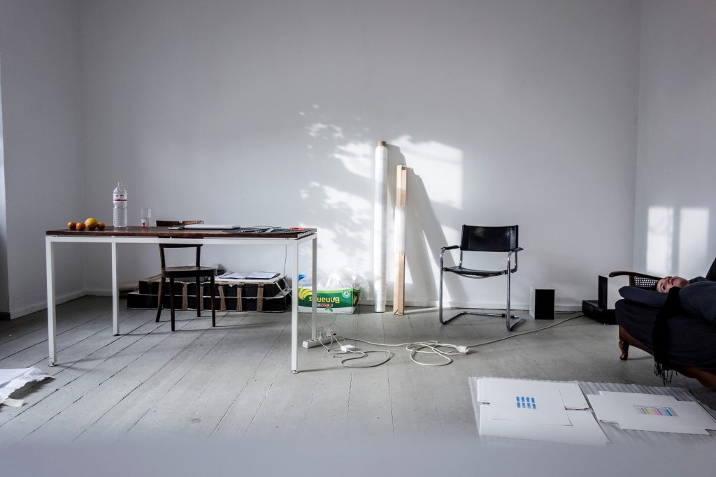 Ein weißgestrichener Raum mit hellem Boden, links hinten ein Schreibtisch mit Stuhl, rechts ein Sofa mit einer darauf augestreckten dunkelgekleideten Person. Es ist nur der Oberkörper und das Gesicht zu sehen.