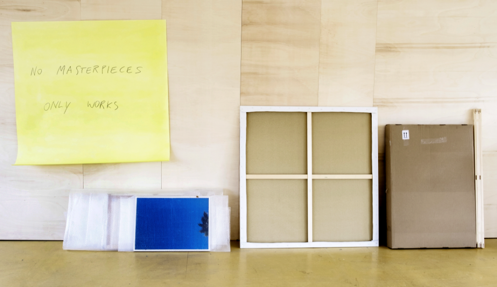 Eine Leinwand in Rückansicht sowie diverse andere Objekt vor einer Holzwand, links ein überdimensionaler gelber Klebezettel in der Größe der Leinwand mit dem Titel der Installation.