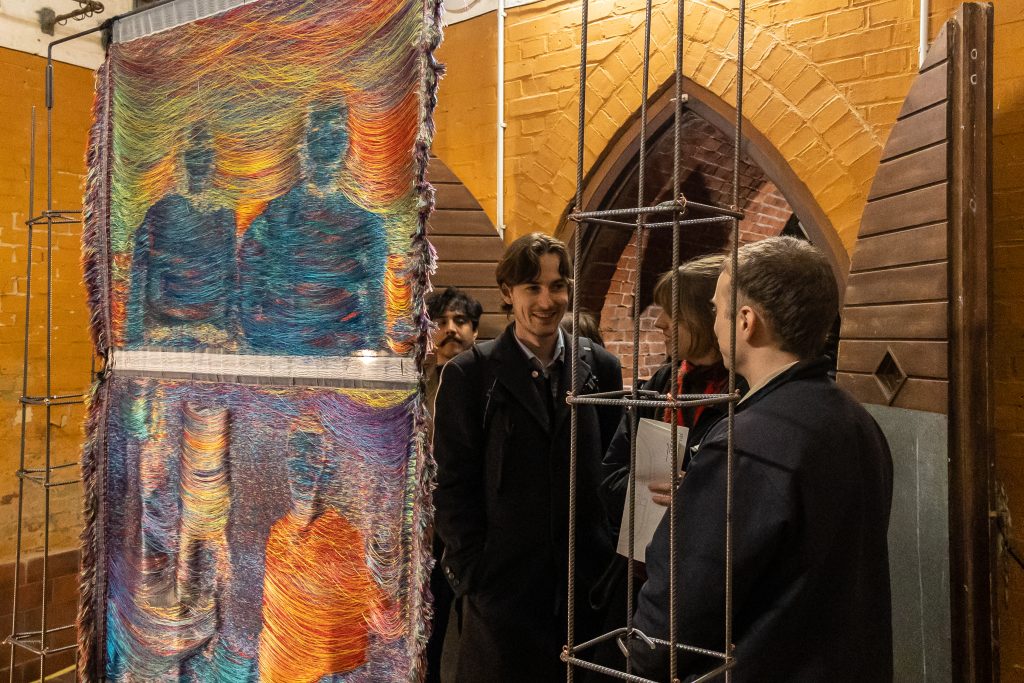 Ein ockerfarben gestrichener Raum, im Hintergrund ist in Spitzbogenportal zu sehen, im Vordergrund ein buntes textiles Kunstwerk und dahinter mehrere Menschen, die sich angeregt unterhalten.