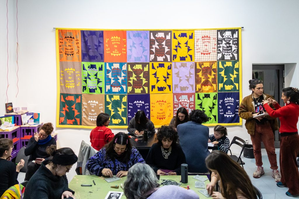 Menschen sitzen an Tischen vor einem bunten Wandbehang und basteln