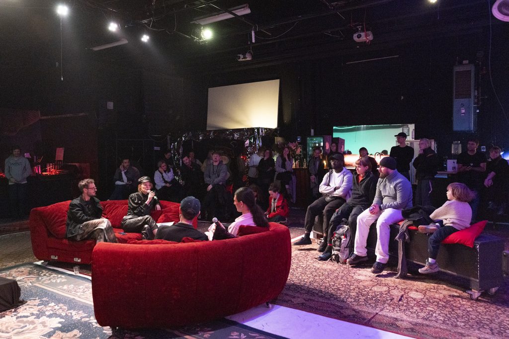 Ein dunkler Studioraum, im Vordergund zwei rote Sofas mit Menschen, die vor Publikum diskutieren, im Hintergrund eine Bar.
