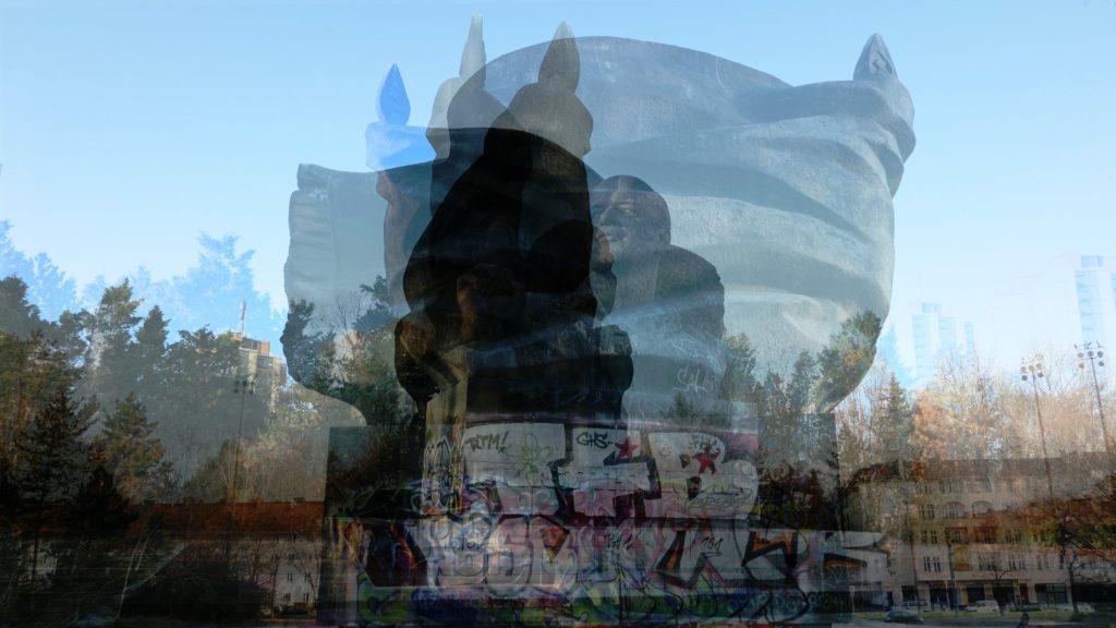 Zu sehen ist ein Standbild, das eine vielfach mit sich selbst überblendete Bronzeplastik eines Mannes vor einer Fahne zeigt. Die Konturen verschwimmen.