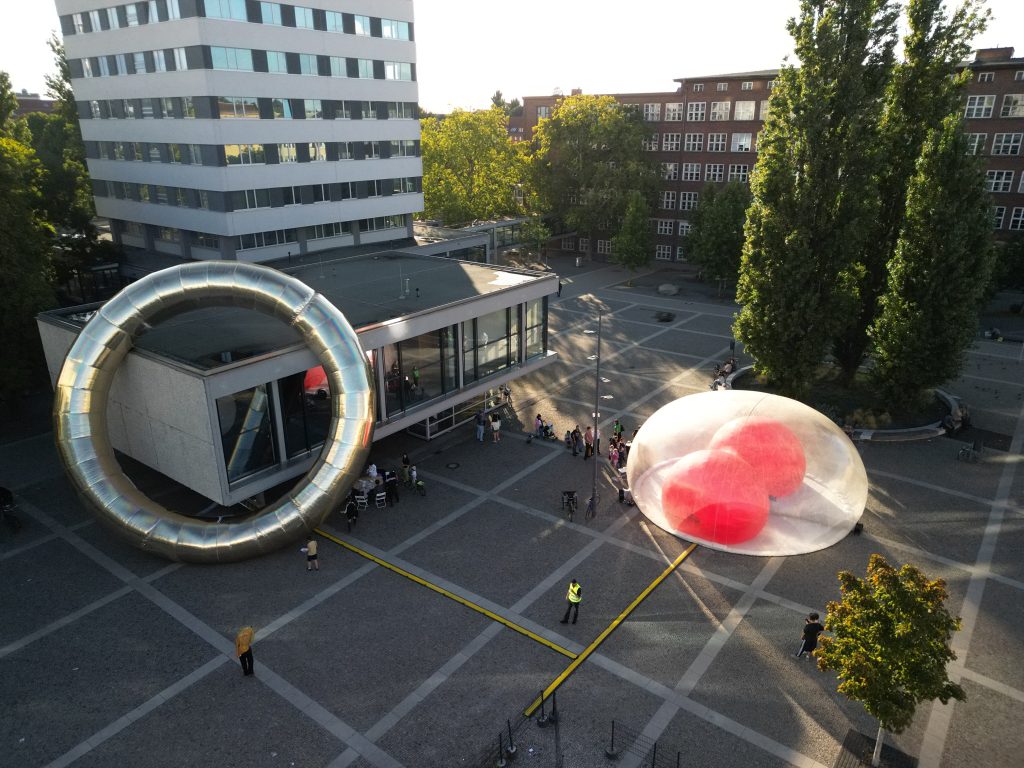 Ein Stdatplatz, links im Bild ein Hochhaus, rechts Bäume, in der Mitte des Bildes eine Installation, bestehend aus einem übergroßen Rind, der sich um ein zweigeschossiges Gebäude legt, sowie einer transparenten Plastikkuppel mit roten Blasen im Inneren.