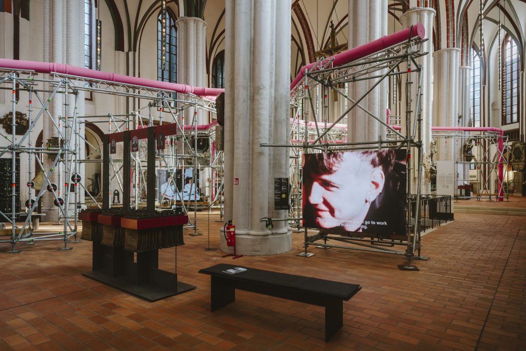 Blick in eine dreischiffige gotische Kirche durch die sich eien Instalattion mit rosafarbenen Rohrleitungen, an den Gerüsten befinden sich diverse Bildschirme.