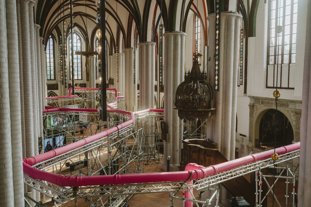 Aus halbhoher Position geht der Blick ins Mittelschiff einer dreischiffigen gotischen Hallenkirche. Zwischen den Säulen schlängeln sich rosafarbene Rohre, die auf einem Metallgerüst montiert sind.