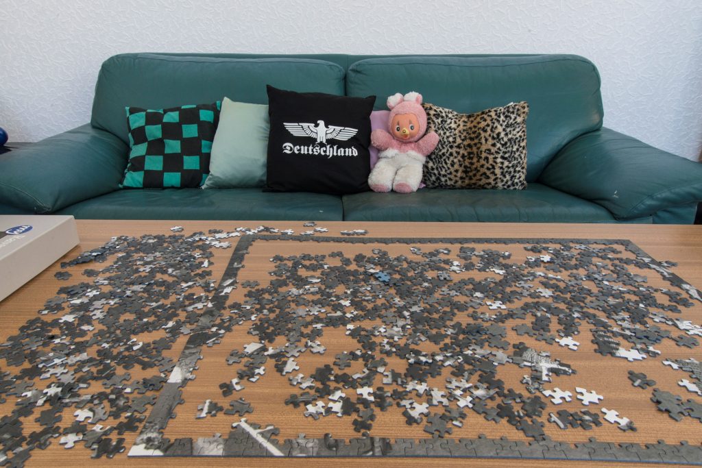 Im Vordergrund auf einem Holztisch ein halbfertiges Puzzle, dahinter ein grünes Sofa mit Kissen und Plüschtier.